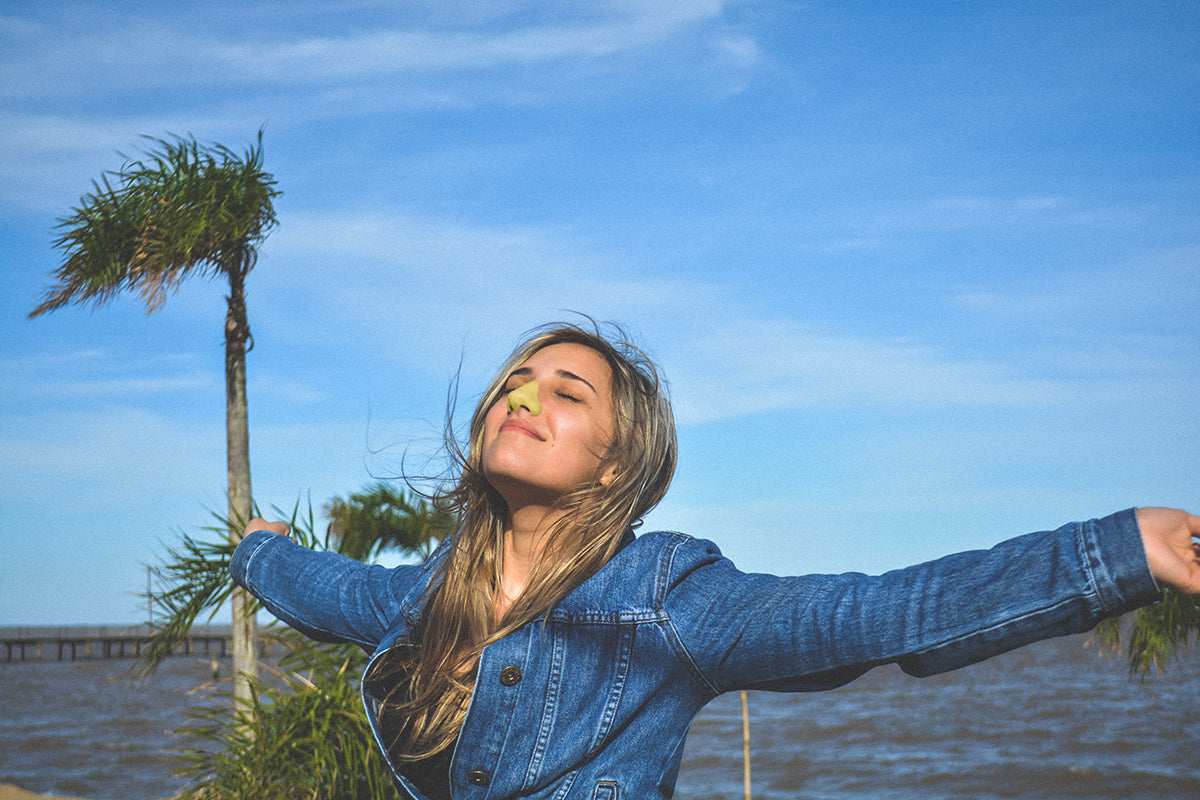 Woman smiles on beach with arms out while wearing yellow Nöz sunscreen on nose