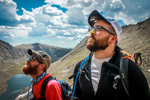 Two men hiking while wearing colorful Nöz SPF in blue and yellow on their noses with mountains in background